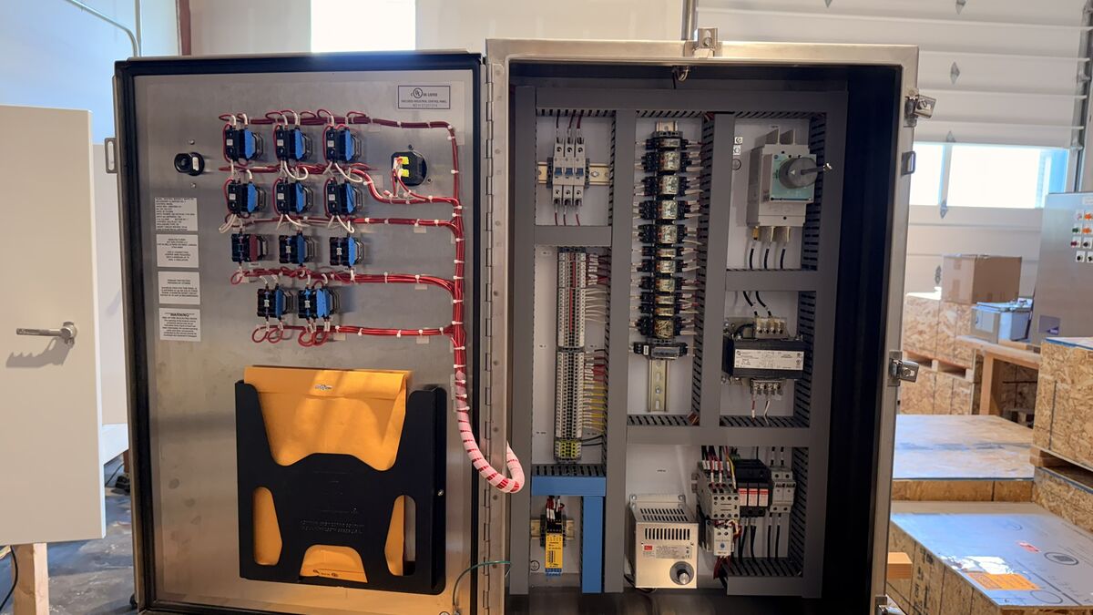 Stainless steel industrial control panel with the door open, showing door-mounted relays with labeled wiring on the left and a subpanel with circuit protection, terminal blocks, a control transformer, and a disconnect switch on the right.