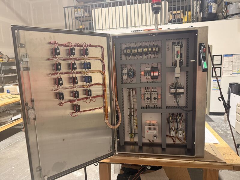 Large stainless control enclosure, door open, showing rows of door-mounted relays with wiring looms and an interior subpanel with a through-door disconnect operator and pilot devices.