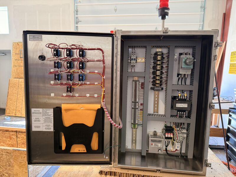 Completed stainless control panel standing open on a wooden pallet in the ACT Solutions shop, with a red stack light on top and a drawing pocket mounted inside the door.