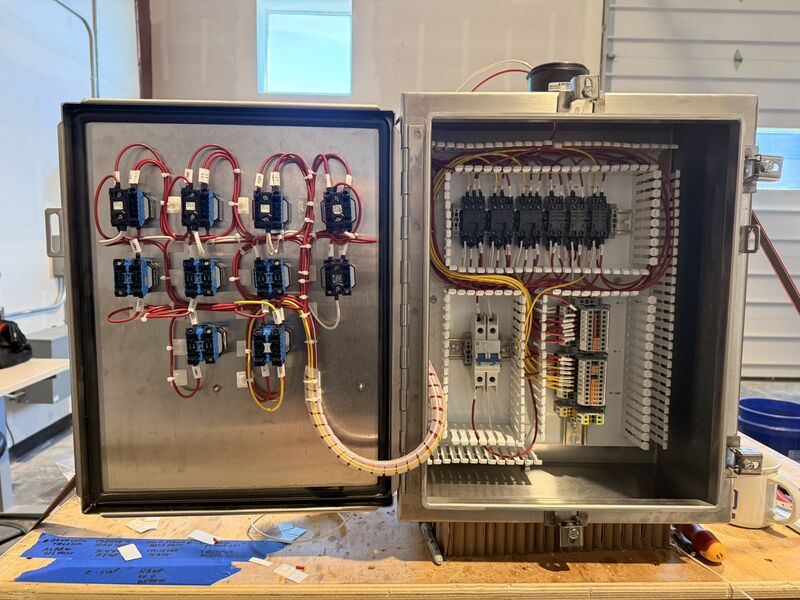 Stainless control panel with the door open on a workbench, relays and labeled wiring mounted to the door and long rows of field terminal blocks on the interior subpanel.