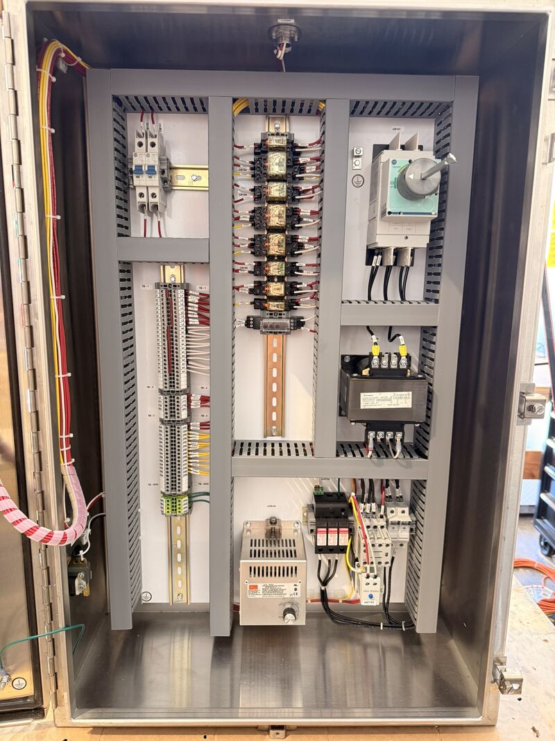 Interior of a control panel showing a vertical column of grey and green terminal blocks, relays on DIN rail, a control transformer, a disconnect switch and a DC power supply, with wire duct routing between them.